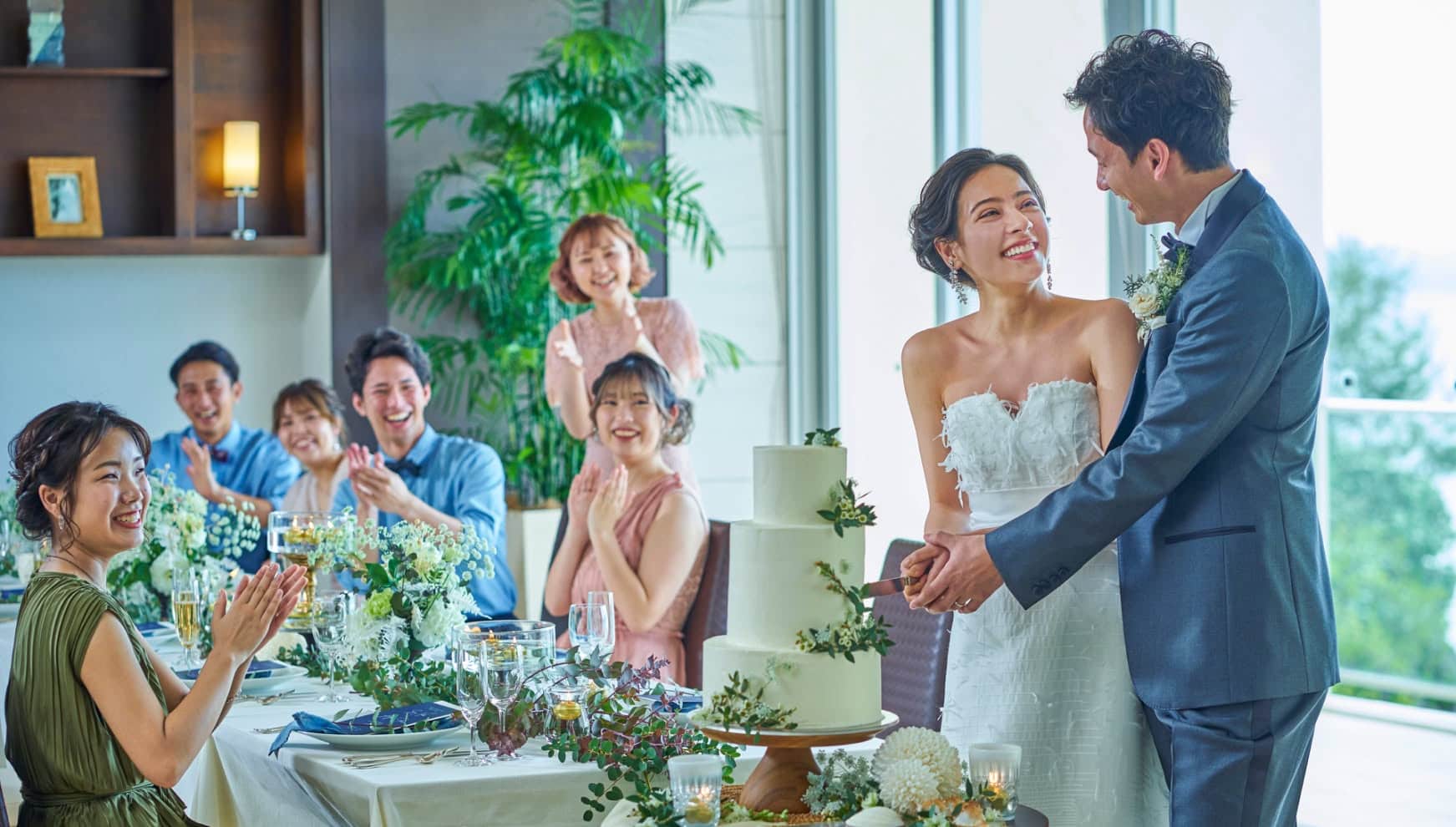 アイネス ヴィラ ノッツェ 沖縄のパーティ会場にて、ゲストの祝福を浴びながらウエディングケーキに入刀する新郎新婦。海が見える明るい会場で、ゲストが笑顔で拍手を送るアットホームで幸せ溢れるケーキカットのシーン。