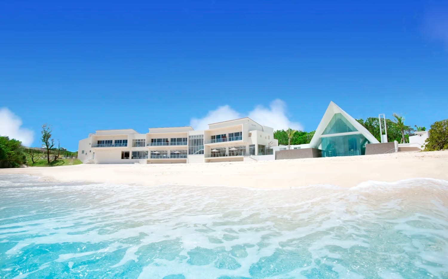 沖縄の名護に位置するアイネス ヴィラ ノッツェ 沖縄の全景。青い空と澄んだ海、白い砂浜が広がるビーチフロントに、三角形の特徴的なチャペルとモダンなヴィラが並ぶ、リゾート感あふれるウエディングステージの風景。