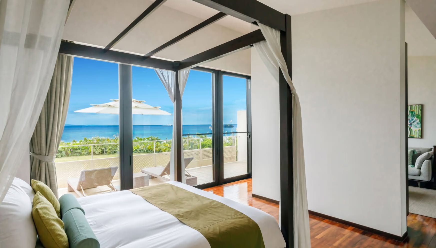 アイネス ヴィラ ノッツェ 沖縄のオーシャンビュー客室。天蓋付きのキングサイズベッドが備えられ、大きな窓からは沖縄の青い海とテラスのビーチチェアが見渡せる。白と木目を基調とした清潔感あふれる贅沢な宿泊空間。