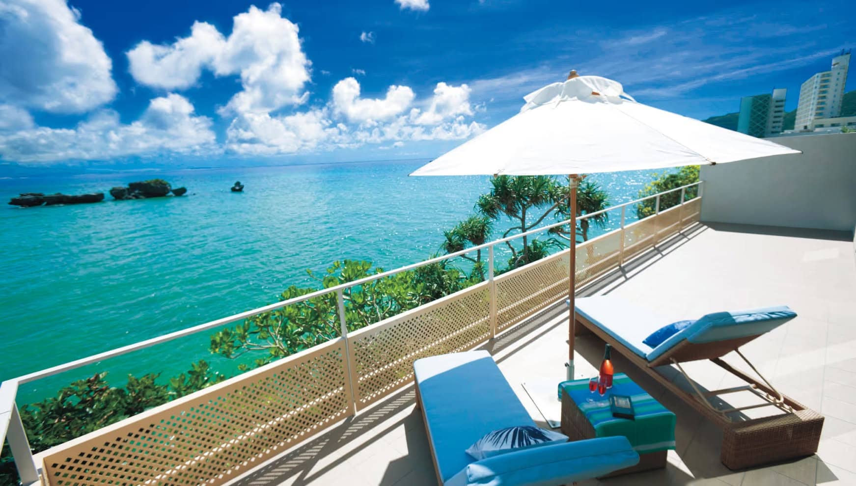 アイネス ヴィラ ノッツェ 沖縄の広々としたヴィラテラス。白いパラソルの下、青いクッションのビーチチェアに横たわり、エメラルドグリーンの美しい海と広大な青空を眺めながら過ごす、至福のリゾートウエディングのひととき。