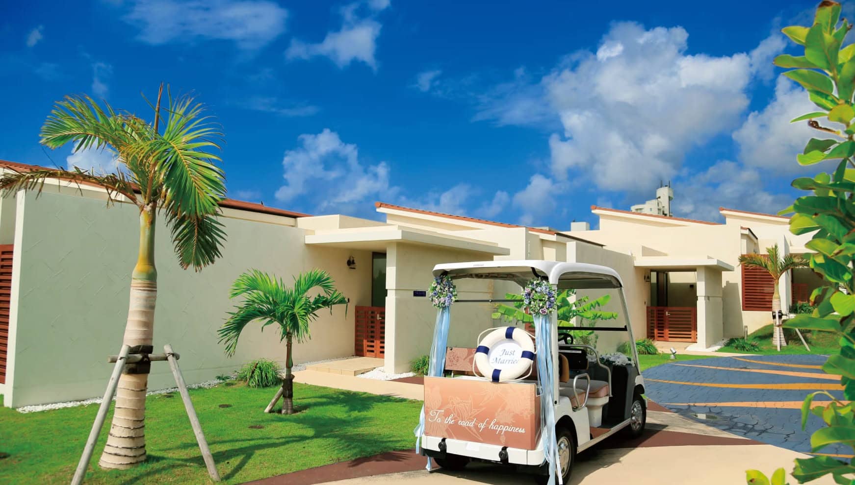 アイネス ヴィラ ノッツェ 沖縄の敷地内を移動するウエディング仕様のカート。背面に「Just Married」のロゴと浮き輪の装飾が施され、青い空とヤシの木、そしてモダンなヴィラが並ぶリゾート感溢れる風景。