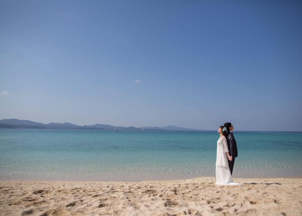 アイネス ヴィラ ノッツェ 沖縄の目の前に広がる天然ビーチにて、背中合わせに立ち手をつなぐ新郎新婦。澄み渡る青い空とエメラルドグリーンの海を背景に、穏やかな波打ち際で静かに目を閉じ、リゾートの風を感じる幻想的なウエディングフォト。