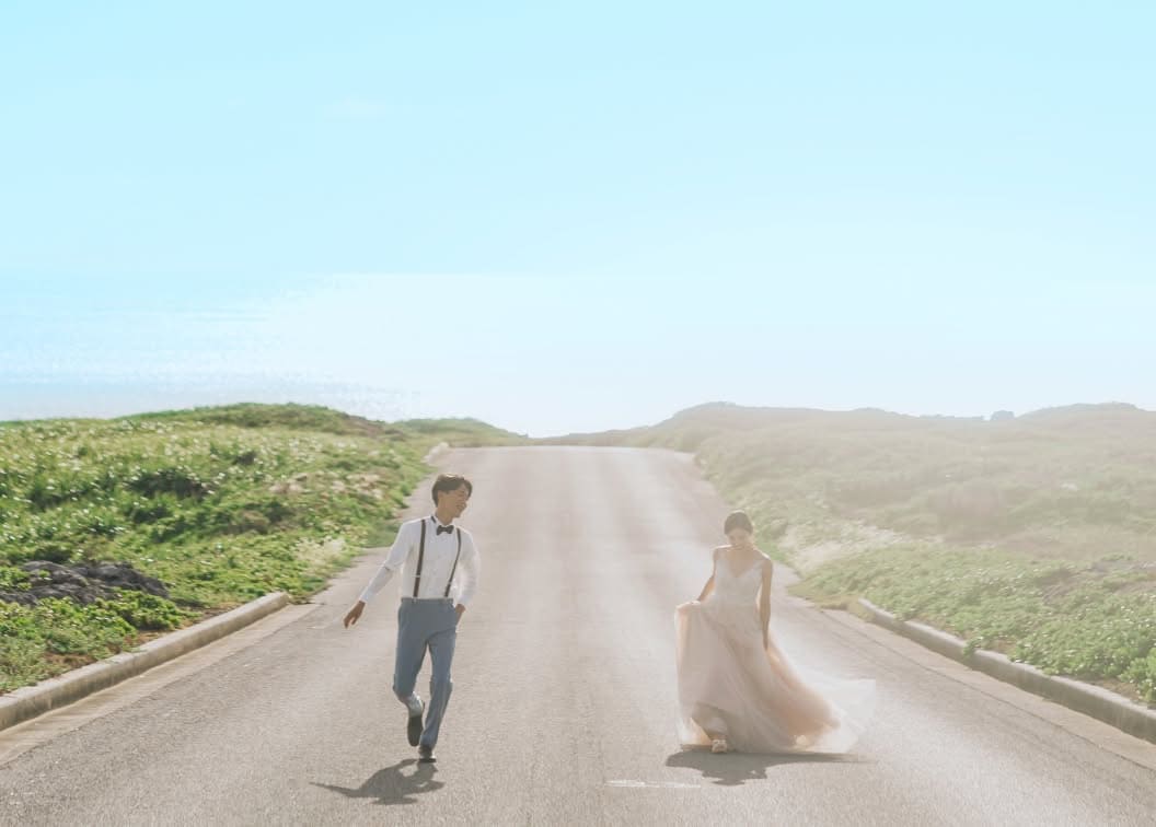 アイネス ヴィラ ノッツェ 沖縄の周辺、海へと続く一本道を歩く新郎新婦。柔らかな日差しが二人を包み込み、ウエディングドレスの裾を軽やかに持ち上げて歩く新婦と、笑顔で寄り添う新郎の幸せなロードムービーのような一景。