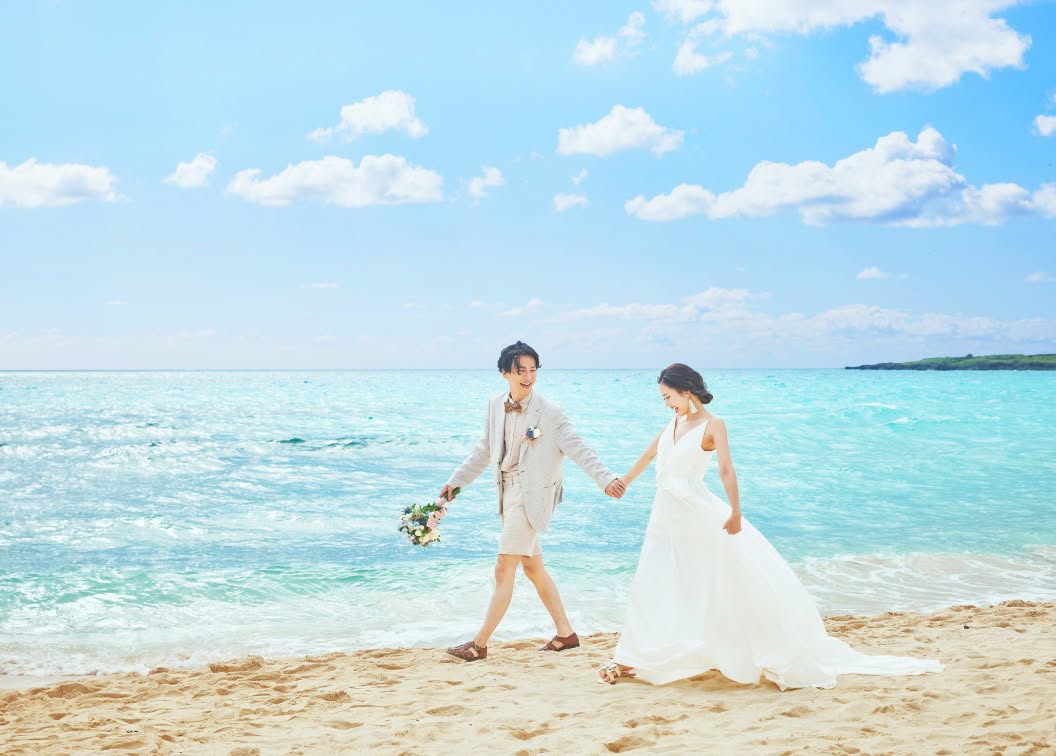 アイネス ヴィラ ノッツェ 沖縄の目の前に広がる白砂のビーチを、手をつないで歩く新郎新婦。エメラルドグリーンの澄んだ海とどこまでも続く青い空を背景に、ウエディングドレスとハーフパンツスタイルの新郎が笑顔を見せる開放的なロケーションフォト。