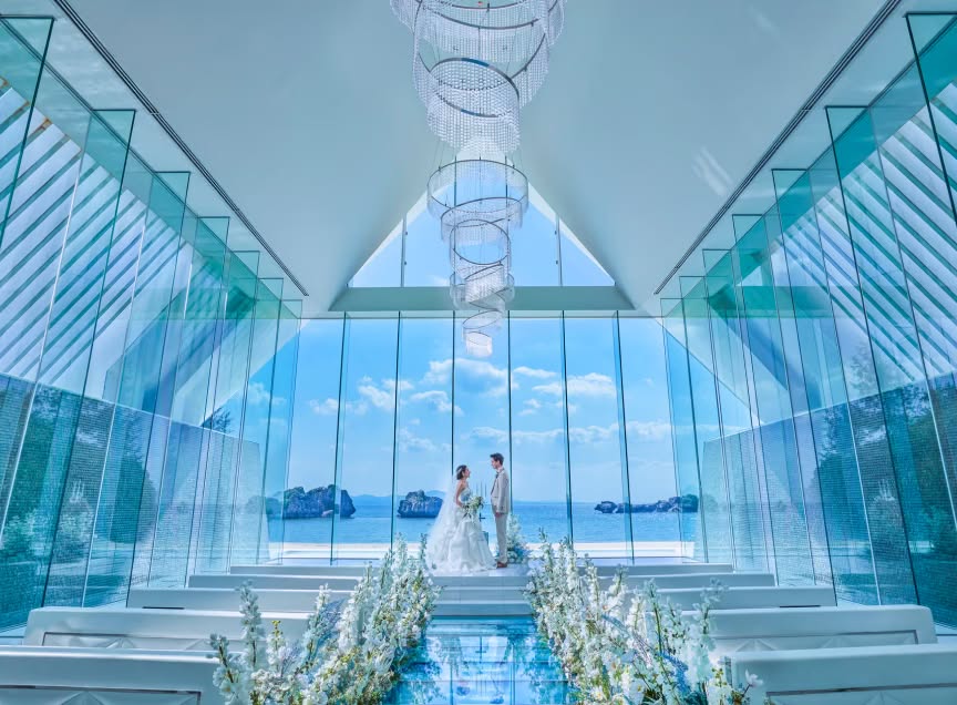 アイネス ヴィラ ノッツェ 沖縄の全面ガラス張りのチャペルにて、海を背景に見つめ合う新郎新婦。純白のドレスとタキシード姿の二人が、高い天井から吊るされたシャンデリアと白い花々に囲まれた幻想的な空間に立つ。