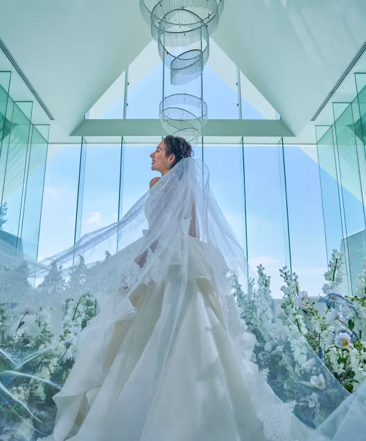 アイネス ヴィラ ノッツェ 沖縄のチャペル。ガラス張りの空間で、新婦のロングベールが風になびき、幻想的に舞い上がる。足元は白い花々に囲まれ、背景の青空と光がウエディングドレスを際立たせる、神秘的な一枚。