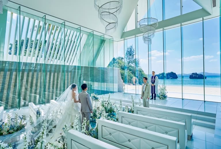 アイネス ヴィラ ノッツェ 沖縄の透明感溢れるチャペルにて、祭壇へ向かう新郎新婦。全面ガラス張りの向こうに広がる青い海と、バージンロードを彩る白い花々、そして二人を待つ牧師。光が差し込む神聖なウエディングの瞬間。