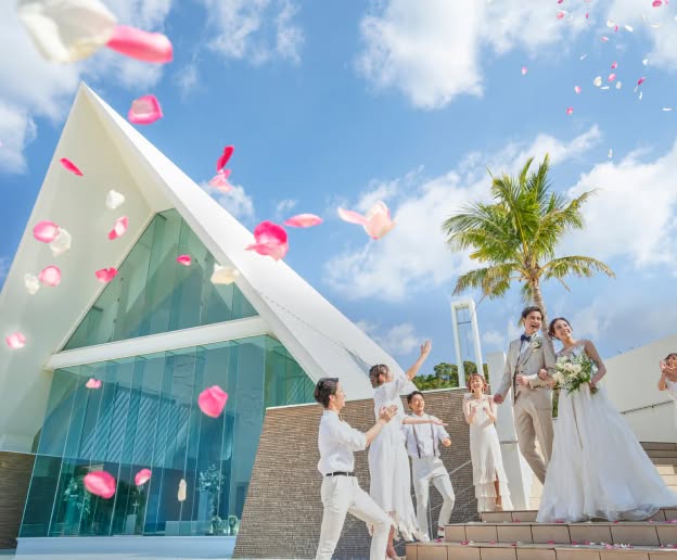 アイネス ヴィラ ノッツェ 沖縄のチャペル外階段にて、ゲストに祝福される新郎新婦。青い空とヤシの木を背に、舞い上がる鮮やかなピンクの花びらの中を笑顔で進む、タキシード姿の新郎とウエディングドレス姿の新婦。