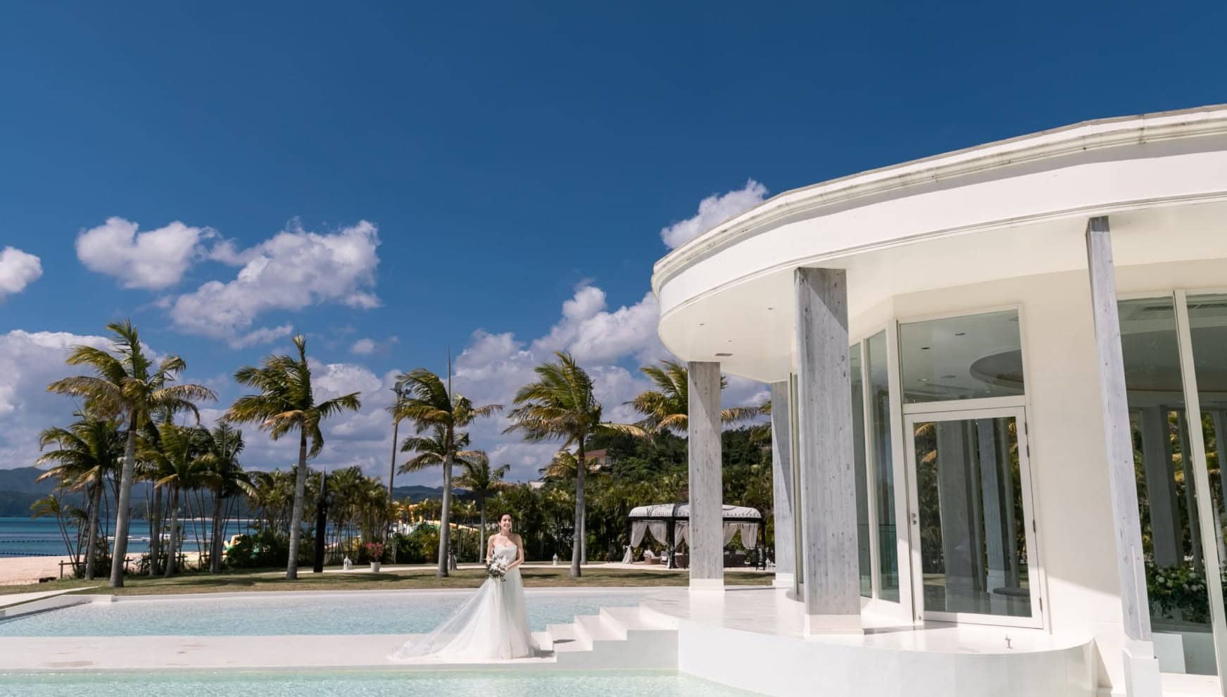 青空の下、パームツリーに囲まれた白いチャペルのプールサイドに、ウエディングドレス姿の新婦が一人佇む。リゾート感あふれるロケーション。