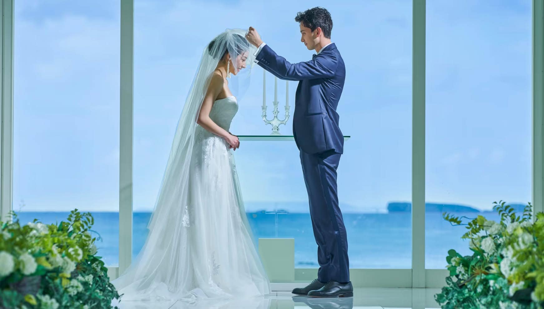 沖縄のカヌチャベイにあるチャペルにて、青い海を背景にベールアップを行う新郎新婦。光が差し込むガラス張りの祭壇前で、タキシードとウエディングドレス姿の二人が誓いを交わす感動的なシーン。