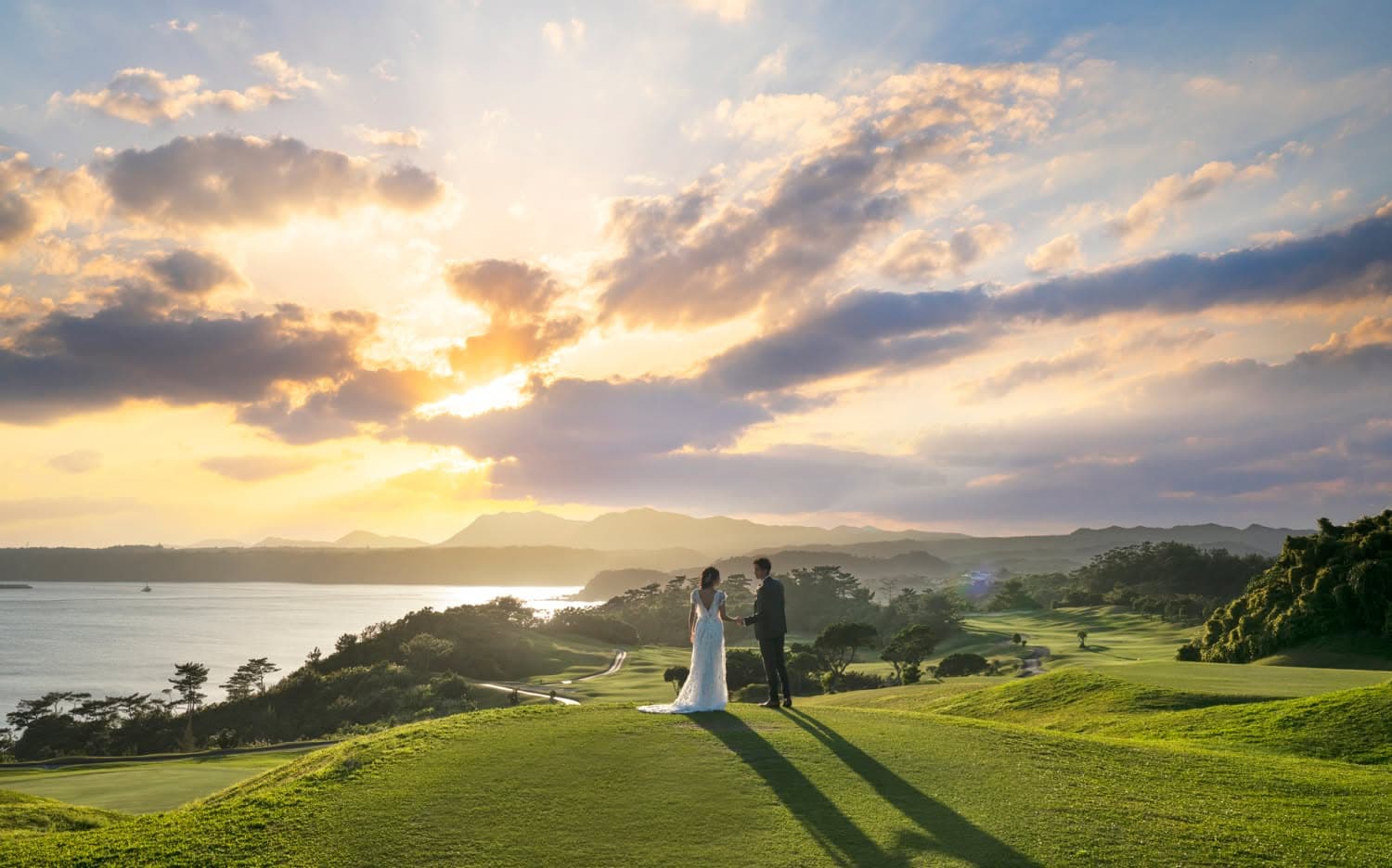 夕暮れ時の沖縄、カヌチャベイの広大なゴルフコースの丘に立つ新郎新婦。黄金色のサンセットと雲間から差し込む光を背景に、タキシードとウエディングドレス姿の二人が手を取り合う、幻想的でロマンチックなロケーションフォト。