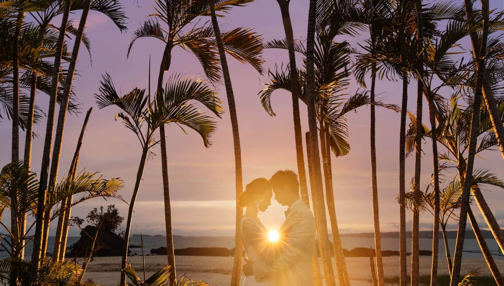 夕暮れ時の沖縄、カヌチャベイのビーチに並ぶヤシの木の間で寄り添う新郎新婦。沈みゆく太陽の光が二人の間で輝き、紫がかった美しい夕空を背景にした幻想的でロマンチックなウエディングフォト。
