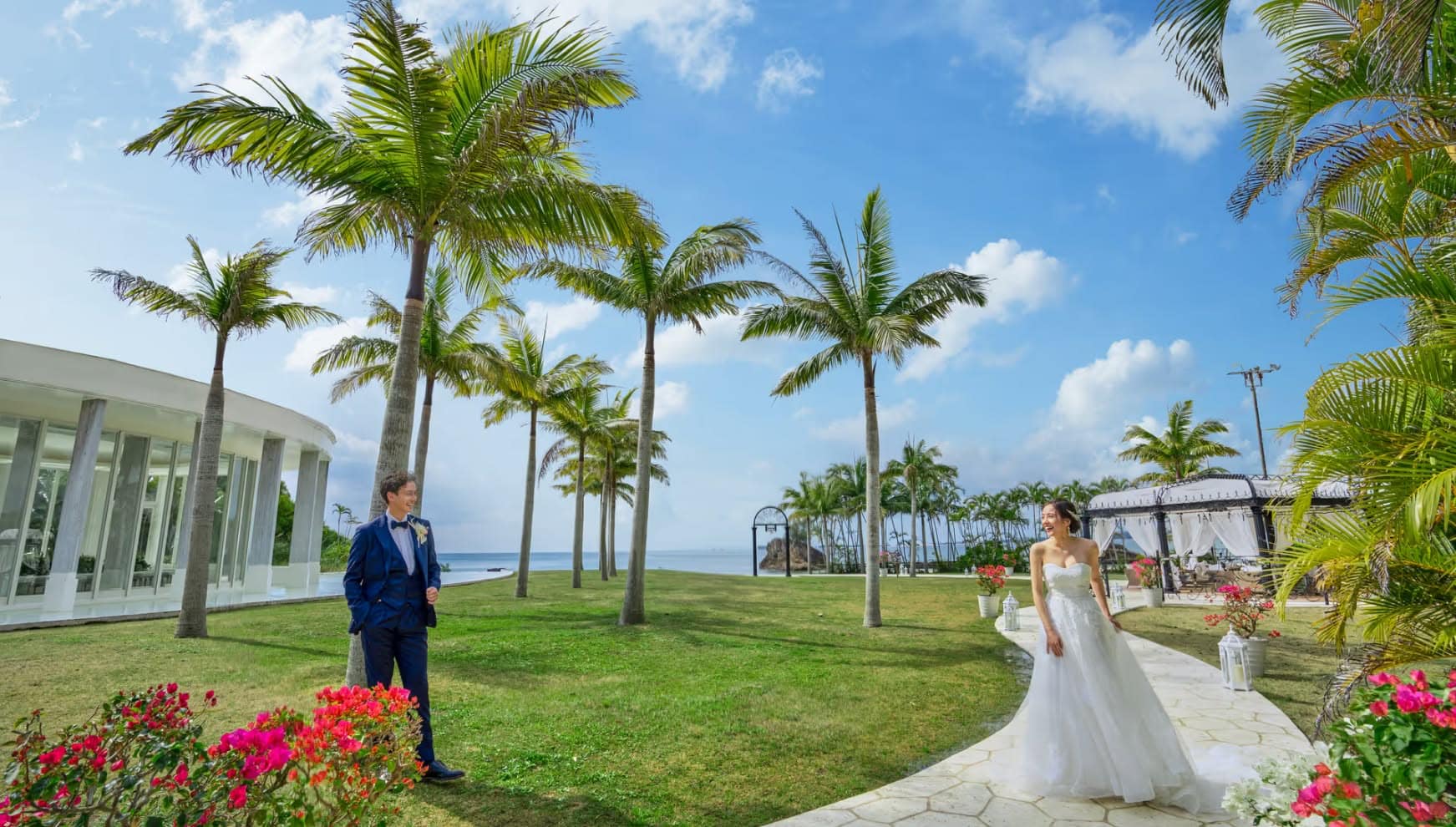 沖縄のカヌチャベイにあるガーデン。青空とヤシの木に囲まれた白い石畳の小道を、笑顔で見つめ合う新郎新婦が歩く様子。タキシードとウエディングドレスが南国の緑に映える爽やかな一枚。