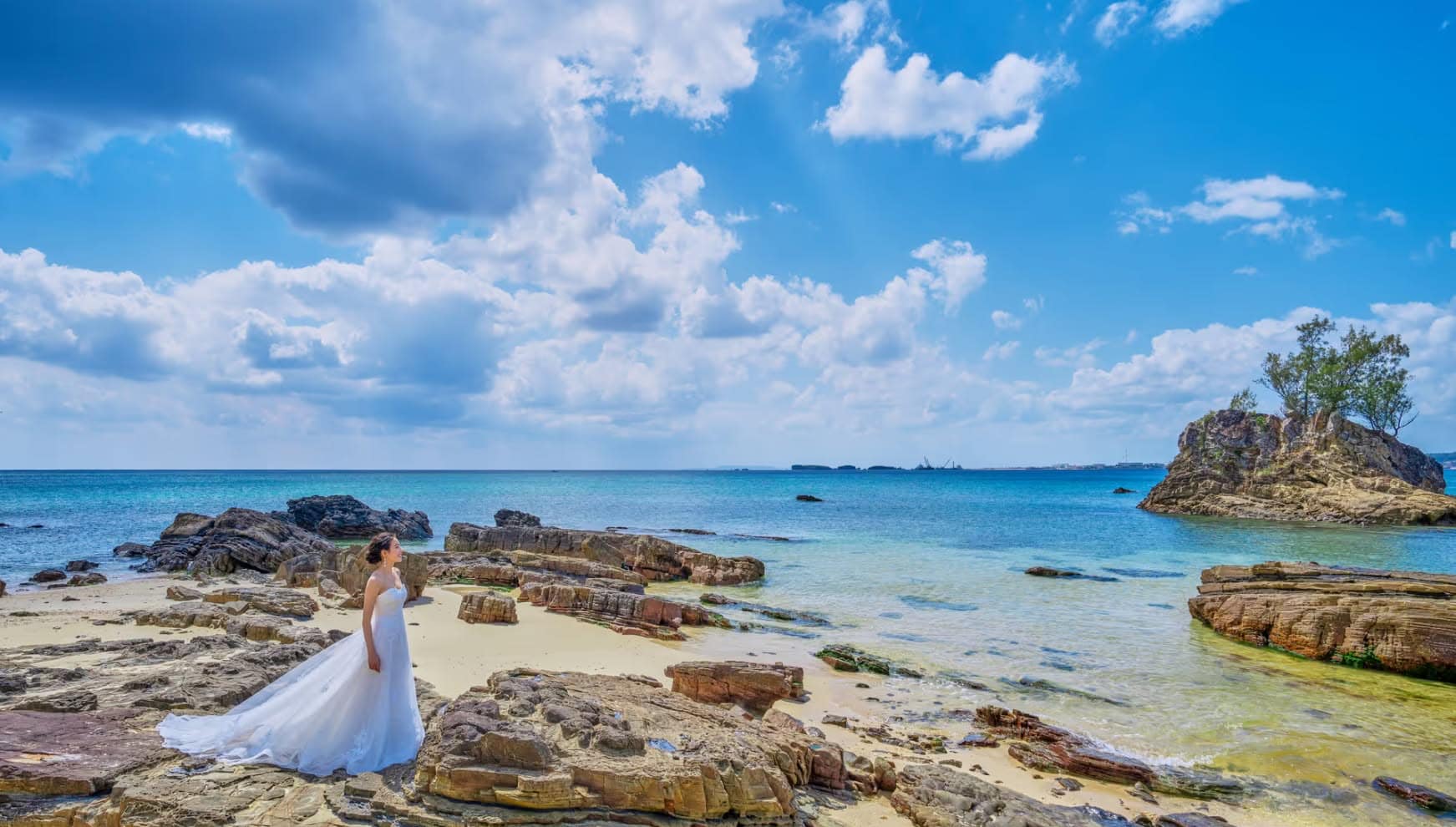 沖縄のカヌチャベイにあるビーチ。エメラルドグリーンの海と雄大な岩場を背景に、純白のウエディングドレスを纏った新婦が一人佇む。自然の造形美が際立つドラマチックなロケーションフォト。