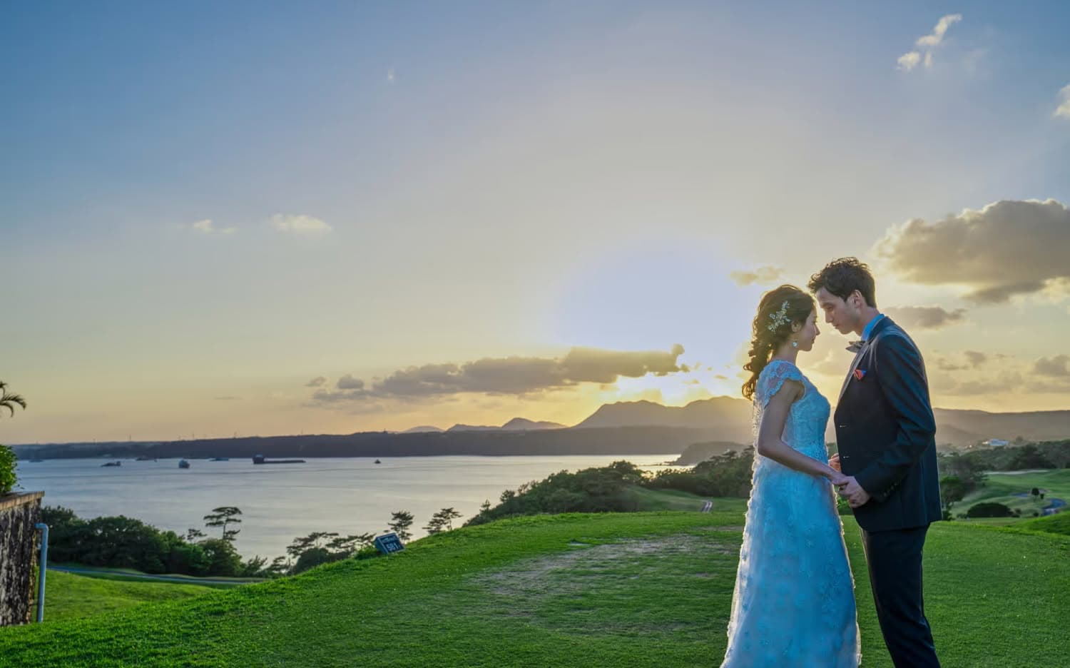 夕暮れ時の沖縄、カヌチャベイの丘の上で向き合う新郎新婦。レースが美しいウエディングドレスとタキシードを纏い、サンセットの柔らかな光に包まれながら静かに手を取り合う、ロマンチックな雰囲気のロケーションフォト。