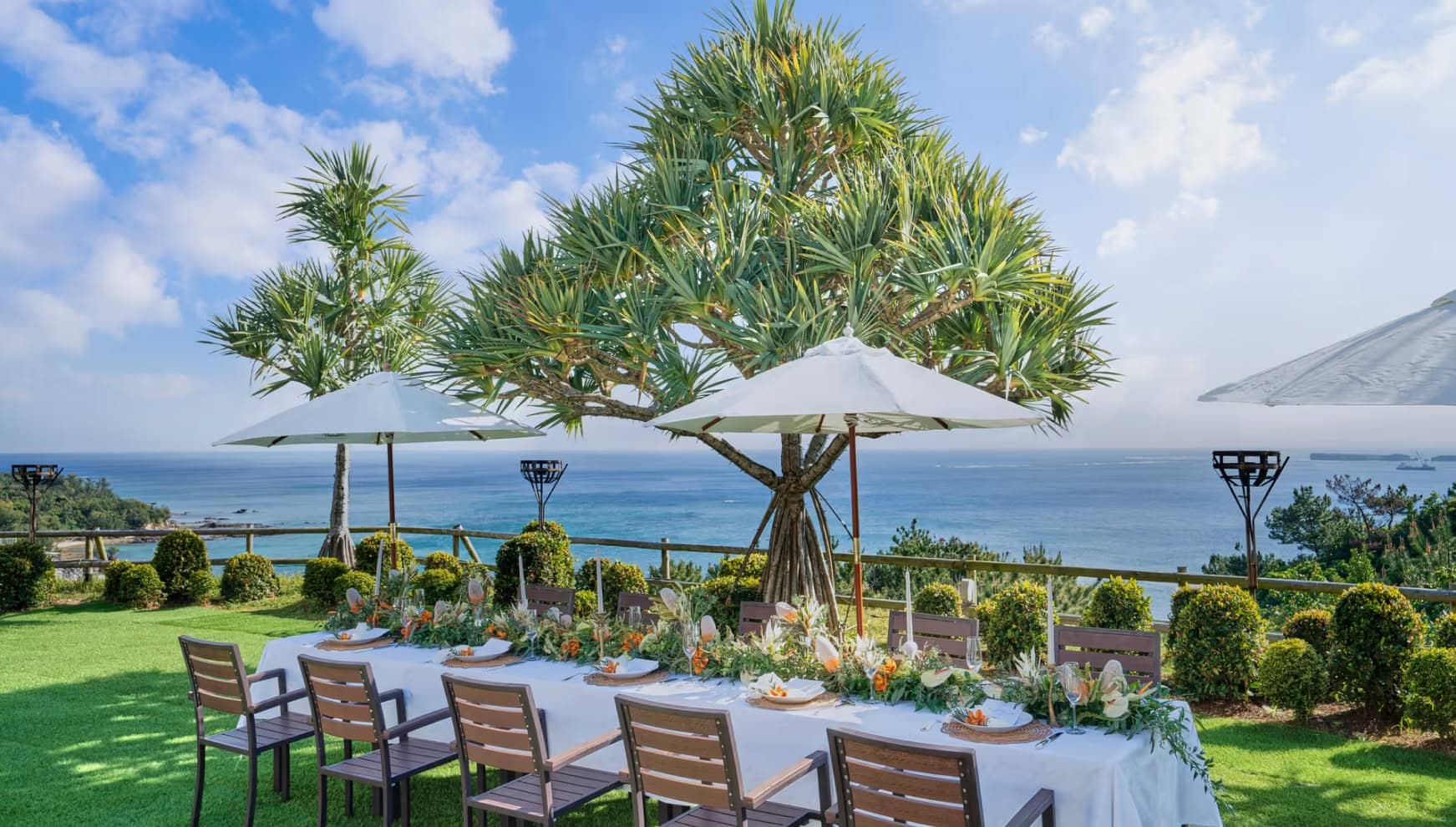 沖縄のカヌチャベイにて、青い海を見渡す芝生の丘に設置された屋外ウエディングのパーティー会場。白いクロスが敷かれた長テーブルに華やかなフラワーアレンジメントが並び、パラソルとヤシの木が南国リゾートを演出する。