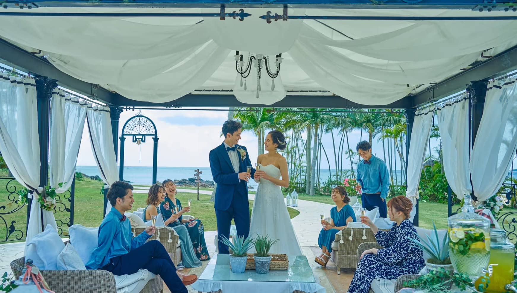 沖縄のカヌチャベイにて、白を基調とした開放的なガゼボで楽しむウエディングパーティー。海を背景に、タキシード姿の新郎とドレス姿の新婦がゲストと笑顔で乾杯を交わす、リラックスしたリゾート宴会の様子。