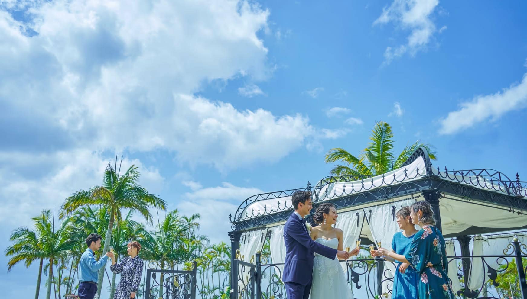 沖縄のカヌチャベイにて、晴れ渡る青空とヤシの木を背景に、装飾が美しいガゼボで乾杯する新郎新婦とゲスト。タキシードとウエディングドレス姿の二人が友人たちと笑顔でグラスを合わせる、開放感あふれるリゾートパーティー。