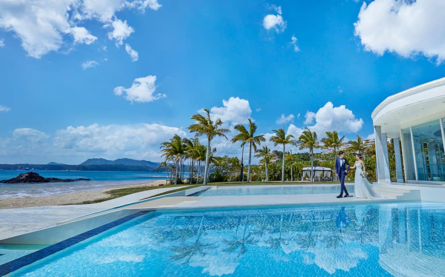 沖縄の名護に位置するアイネス ヴィラ ノッツェ 沖縄の全景。青い空と澄んだ海、白い砂浜が広がるビーチフロントに、三角形の特徴的なチャペルとモダンなヴィラが並ぶ、リゾート感あふれるウエディングステージの風景。