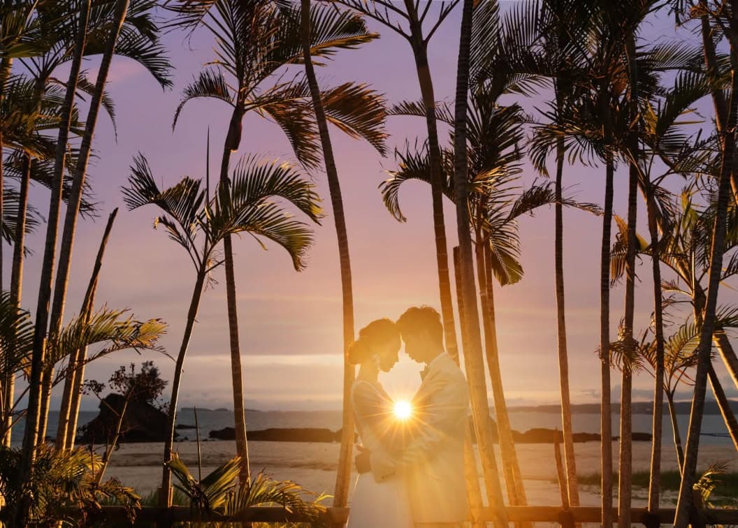 夕暮れ時の沖縄、カヌチャベイのビーチに並ぶヤシの木の間で寄り添う新郎新婦。沈みゆく太陽の光が二人の間で輝き、紫がかった美しい夕空を背景にした幻想的でロマンチックなウエディングフォト。