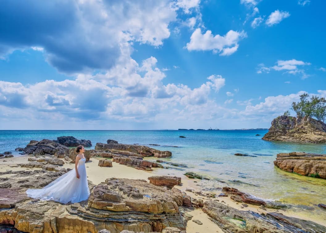 沖縄のカヌチャベイにあるビーチ。エメラルドグリーンの海と雄大な岩場を背景に、純白のウエディングドレスを纏った新婦が一人佇む。自然の造形美が際立つドラマチックなロケーションフォト。