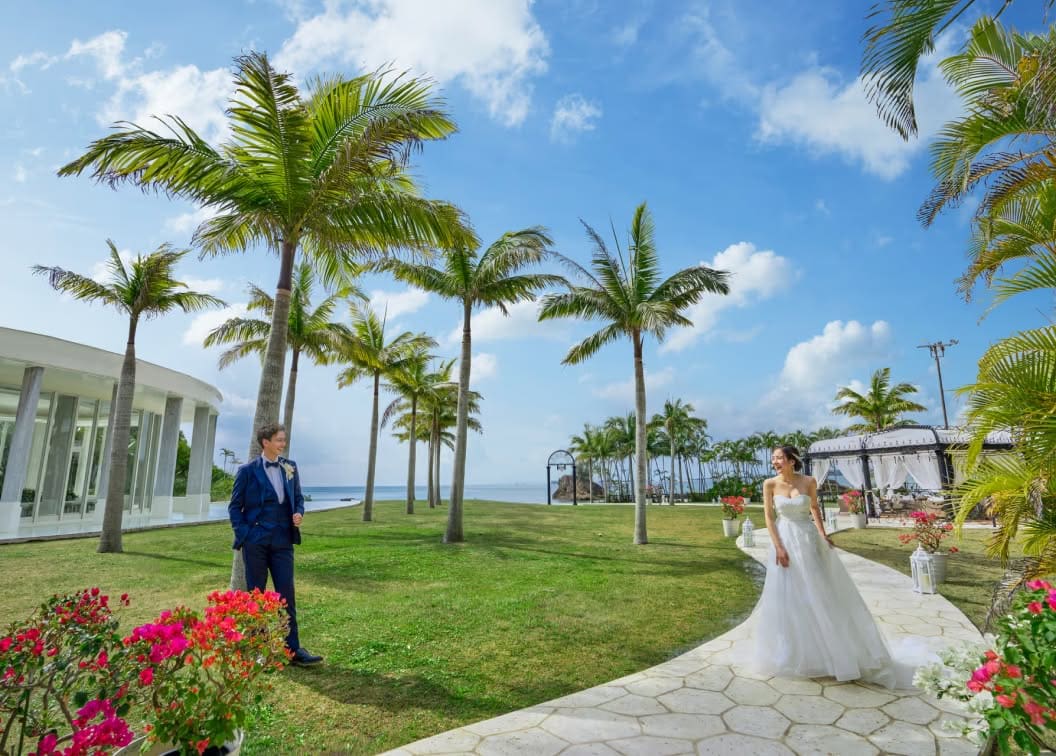沖縄のカヌチャベイにあるガーデン。青空とヤシの木に囲まれた白い石畳の小道を、笑顔で見つめ合う新郎新婦が歩く様子。タキシードとウエディングドレスが南国の緑に映える爽やかな一枚。