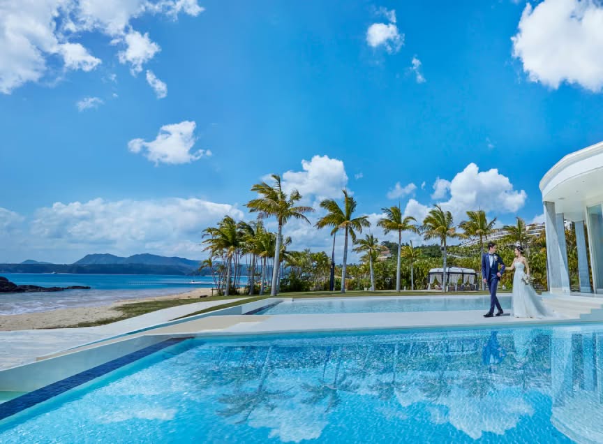 沖縄の名護に位置するアイネス ヴィラ ノッツェ 沖縄の全景。青い空と澄んだ海、白い砂浜が広がるビーチフロントに、三角形の特徴的なチャペルとモダンなヴィラが並ぶ、リゾート感あふれるウエディングステージの風景。