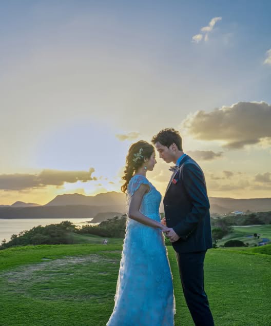夕暮れ時の沖縄、カヌチャベイの丘の上で向き合う新郎新婦。レースが美しいウエディングドレスとタキシードを纏い、サンセットの柔らかな光に包まれながら静かに手を取り合う、ロマンチックな雰囲気のロケーションフォト。