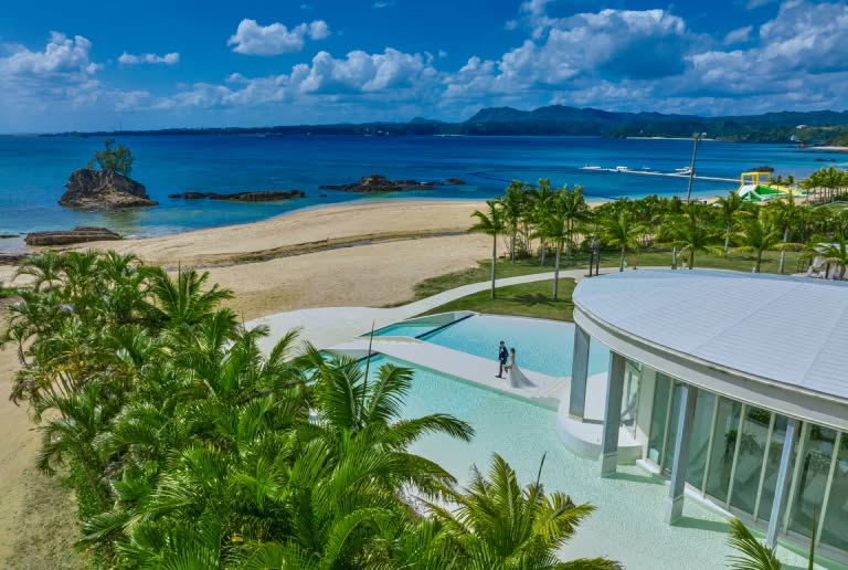 沖縄のカヌチャベイにあるウエディング施設を上空から捉えたカット。青い海と白い砂浜を背景に、ヤシの木に囲まれた水上コテージのようなプールサイドを歩く新郎新婦。南国リゾートらしい開放感に満ちたロケーション。