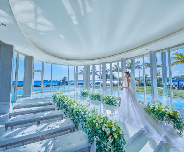 沖縄のカヌチャベイにあるガラス張りのチャペル。青い海と空、ヤシの木を望む開放的な空間で、光が差し込むバージンロードに立つウエディングドレス姿の新婦。白を基調とした洗練された雰囲気。
