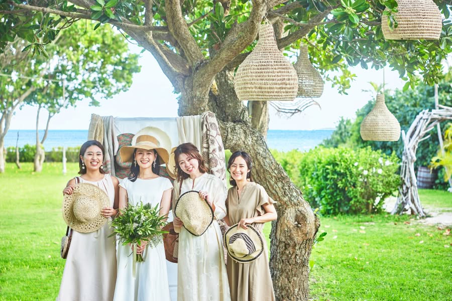 開放的な海を背景にしたガーデンウエディングで、麦わら帽子を手にした友人たちと並び、笑顔を見せるウエディングドレス姿の花嫁。グリーンのブーケを持ち、木々に吊るされたナチュラルな装飾に囲まれた、アットホームで明るい雰囲気の1枚。