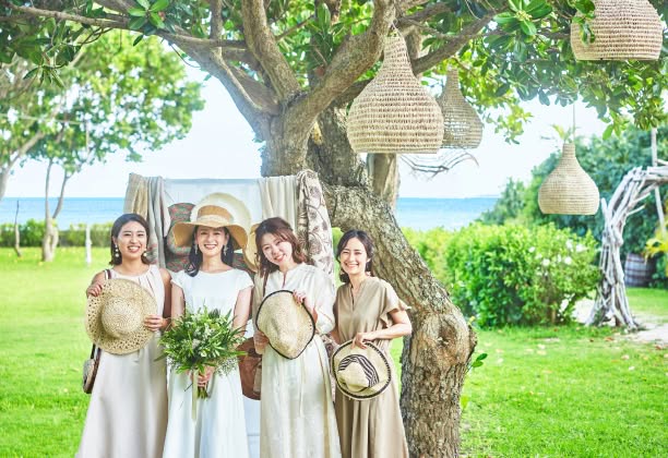 緑豊かな木々の下、海を背景に友人たちと麦わら帽子を手に笑顔を見せる花嫁。ウエディングドレス姿でグリーンのブーケを持ち、ナチュラルな装飾が施されたガーデンウエディング会場で、ゲストとリラックスした時間を楽しむアットホームな1枚。