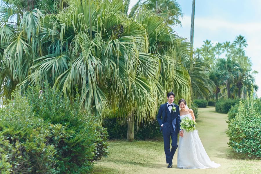 南国風の大きなヤシの木が並ぶガーデンウエディング会場で、手をつないで歩く新郎新婦。ネイビーのタキシードを着た新郎と、白いレースのウエディングドレスを纏いブーケを持った花嫁が、芝生の道を笑顔で進む開放感あふれるシーン。