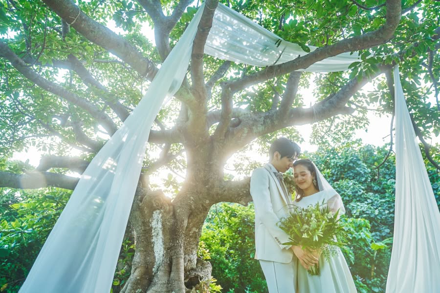 豊かな緑に囲まれたガーデンウエディングで、大きな樹木の下に寄り添い立つ新郎新婦。木漏れ日が差し込む中、白いウエディングドレス姿の花嫁とタキシード姿の新郎が、グリーンのブーケを手に静かに目を閉じている幻想的で温かいシーン。