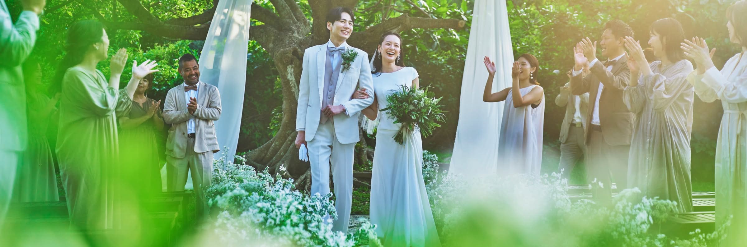 緑豊かな庭園で、ゲストの拍手に包まれながらバージンロードを歩くウエディングドレス姿の花嫁とタキシード姿の新郎。白い花々に囲まれたナチュラルな屋外会場で、腕を組みながら満面の笑みを浮かべる幸せそうな二人の様子。