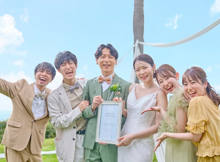 青空の下、ガーデンウエディングの挙式後に結婚証明書を掲げて友人と笑い合う新郎新婦。グリーンのタキシードを着た新郎と白いウエディングドレスの花嫁を囲み、参列した友人たちが満面の笑みを浮かべる、賑やかで幸せ溢れる1枚。