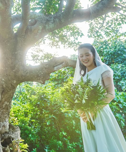 豊かな緑に囲まれたガーデンウエディング会場で、大きな樹木を背にブーケを抱えて微笑む花嫁のソロショット。柔らかな木漏れ日が差し込む中、白いウエディングドレスとベールを纏い、幸せそうな表情を浮かべるナチュラルな雰囲気の1枚。