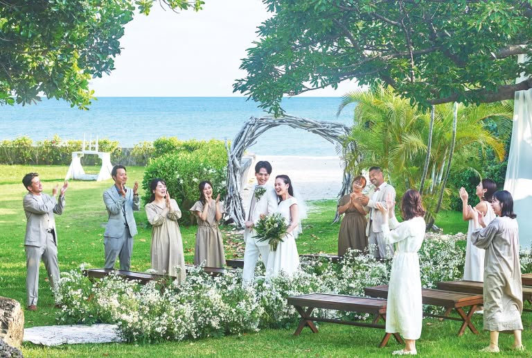 海を一望できる開放的なガーデンウエディング会場で、ゲストの温かな拍手に包まれながら退場する新郎新婦。白いウエディングドレス姿の花嫁と白のタキシードを着た新郎が、笑顔で腕を組み、白い花々に彩られたバージンロードを歩く幸せな光景。