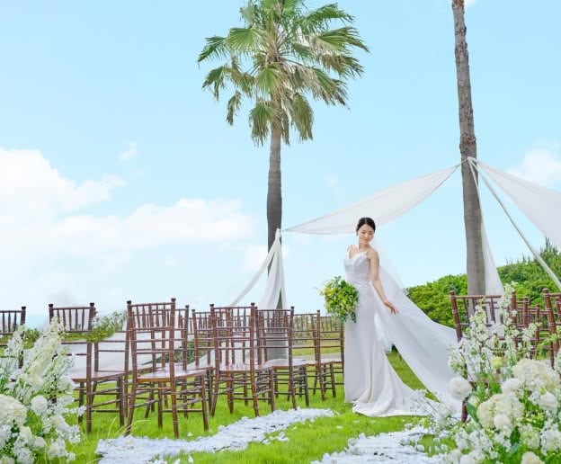 青空と海を望む開放的なガーデンウエディング会場で、パームツリーの下に佇む花嫁。白いウエディングドレスと長いベールを纏い、白い花々に彩られたバージンロードと木製の椅子が並ぶ屋外挙式会場で、静かに微笑む優雅なワンシーン。