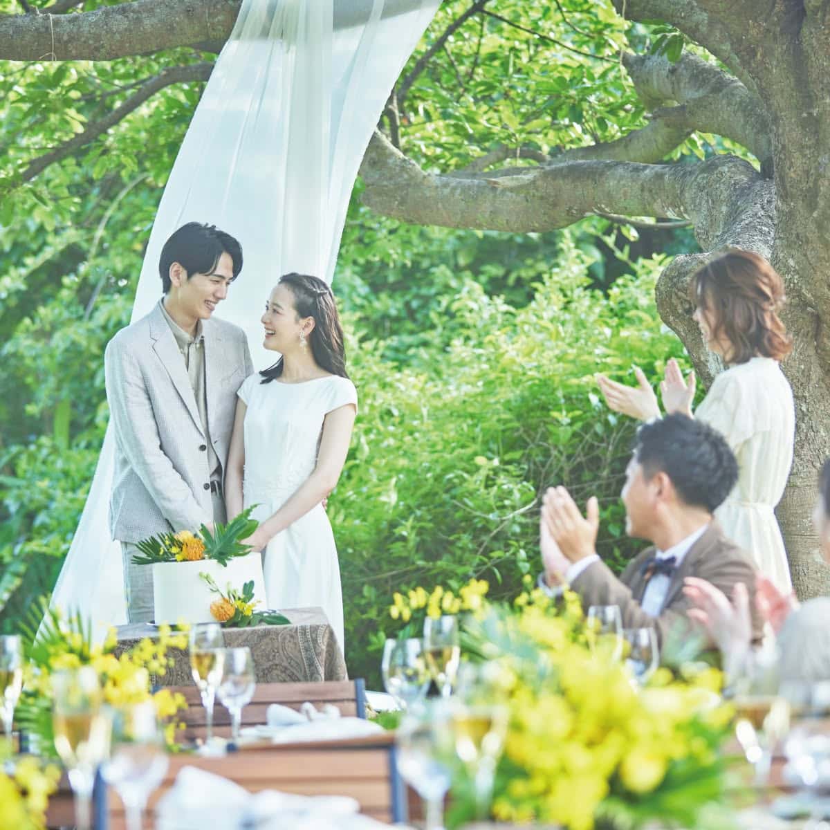 青い空とパームツリーが美しいガーデンウエディング会場で、白い花々に彩られたバージンロードの傍らに立つ花嫁。ウエディングドレスと長いベールを纏い、木製の椅子が並ぶ屋外挙式会場でブーケを手に佇む、開放感あふれるワンシーン。