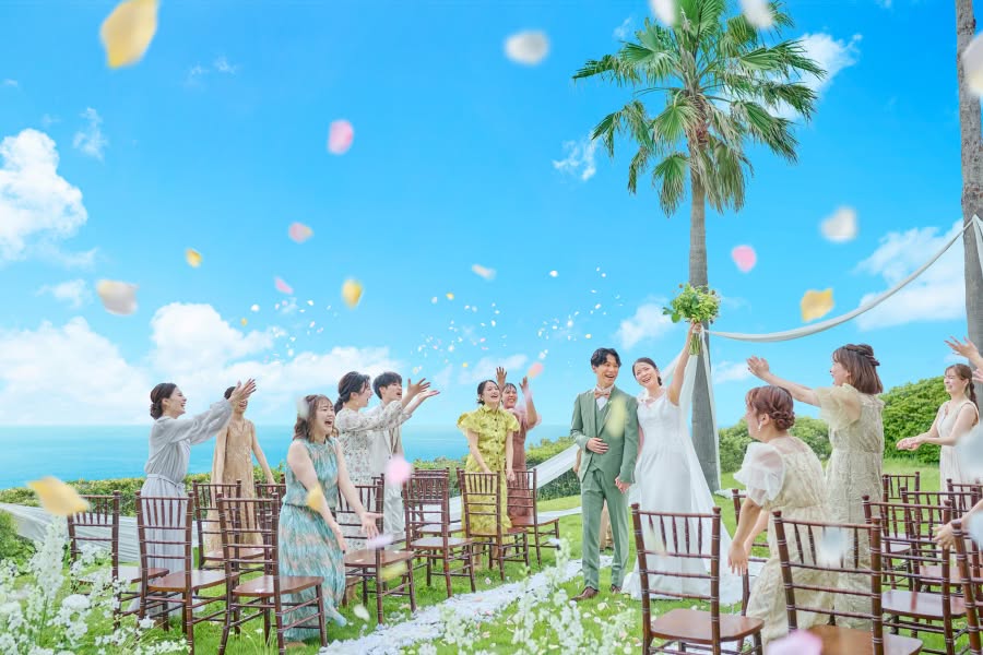 青く澄んだ海と空が広がる絶好のロケーションで行われるガーデンウエディング。フラワーシャワーの祝福の中、ブーケを高く掲げて笑顔を見せる花嫁と、グリーンのスーツを纏った新郎。ゲストの歓声に包まれた、開放感あふれる幸せな瞬間。