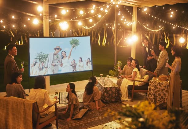 夜のガーデンウエディングで、ライトアップされた屋外会場に設置されたスクリーン映像をゲストと共に楽しむ新郎新婦。温かな電飾の光に包まれたアットホームな雰囲気の中、ソファに座りリラックスして談笑する、ナイトパーティーのひととき。