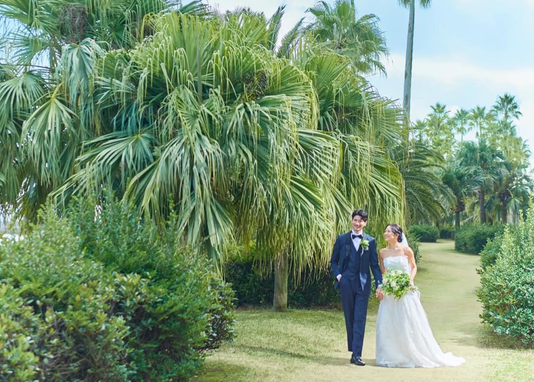 南国風の大きなヤシの木が並ぶガーデンウエディング会場で、手をつないで歩く新郎新婦。ネイビーのタキシードを着た新郎と、白いレースのウエディングドレスを纏いブーケを持った花嫁が、芝生の道を笑顔で進む開放感あふれるシーン。