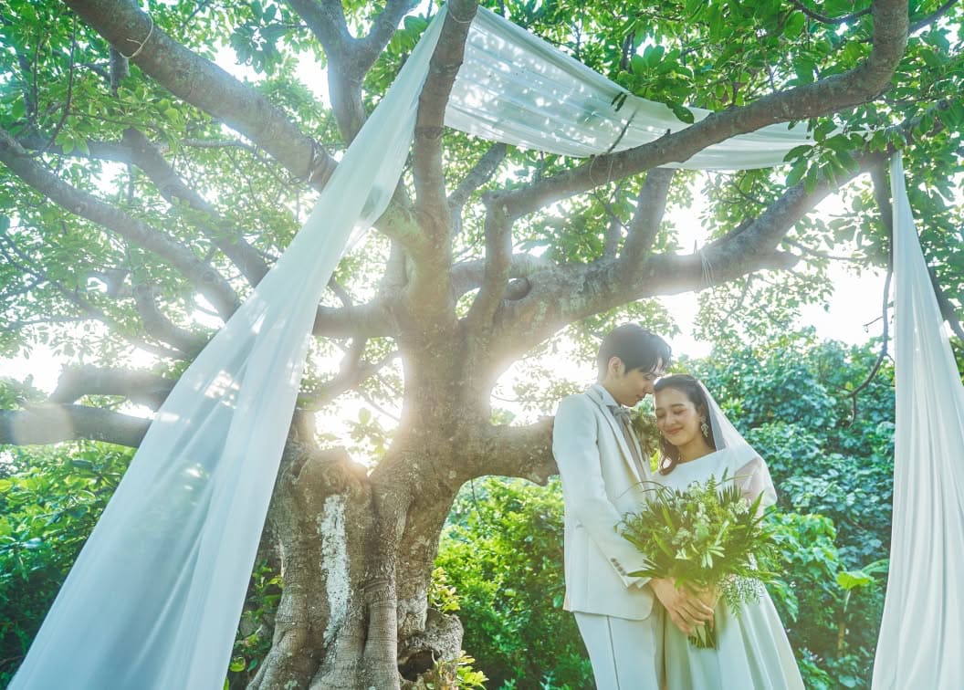 豊かな緑に囲まれたガーデンウエディングで、大きな樹木の下に寄り添い立つ新郎新婦。木漏れ日が差し込む中、白いウエディングドレス姿の花嫁とタキシード姿の新郎が、グリーンのブーケを手に静かに目を閉じている幻想的で温かいシーン。