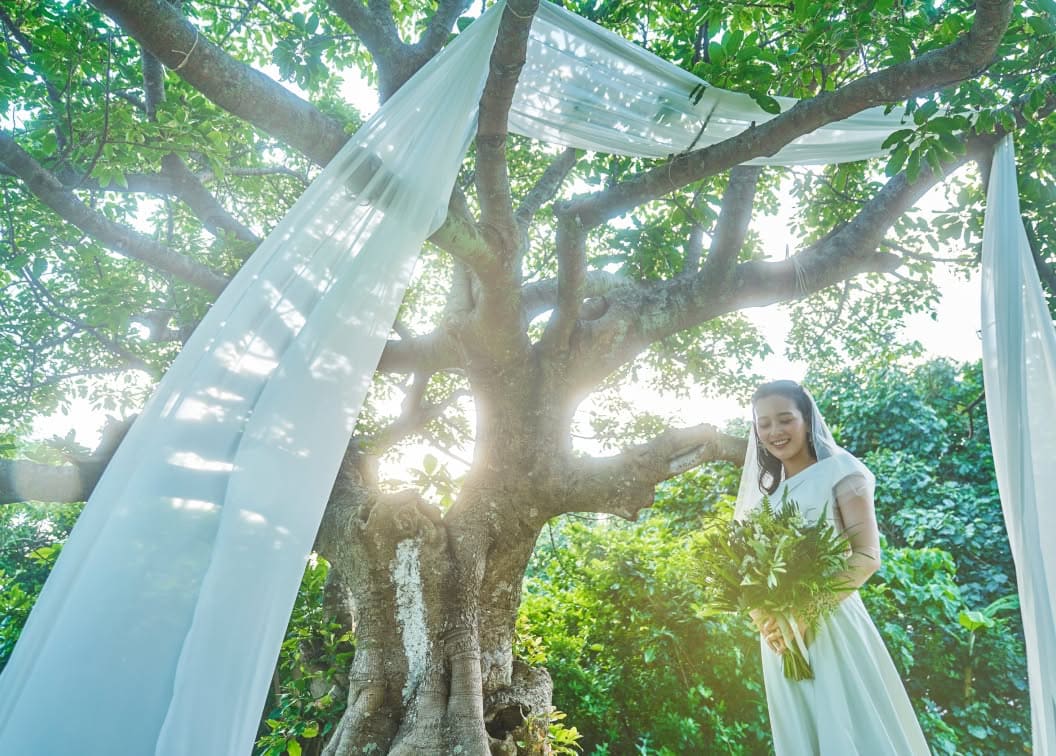 豊かな緑に囲まれたガーデンウエディング会場で、シンボルツリーの傍らに立つ花嫁。白いウエディングドレスとベールを纏い、木漏れ日が差し込む中でグリーンのブーケを手に幸せそうな笑みを浮かべる、ナチュラルで幻想的な雰囲気の1枚。