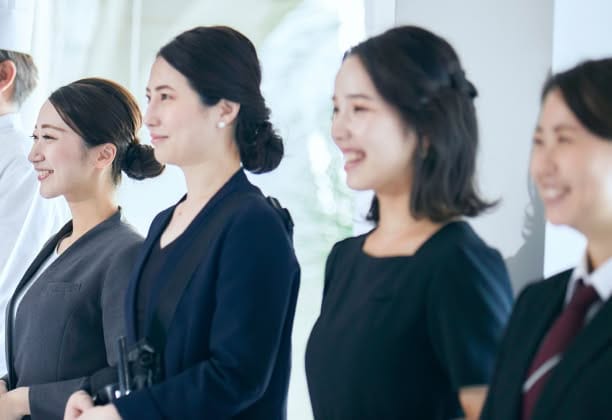 ウエディング会場でゲストを迎えるため、笑顔で一列に並ぶスタッフたちの様子。紺やグレーのスーツを身に纏い、清潔感のある身だしなみで、これから始まるウエディングパーティーに向けて温かく誠実な接客を準備している、信頼感のある表情。