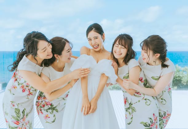 美しい海を背景に、純白のウエディングドレスを着た新婦を囲む4人の女性ゲスト。ゲストはお揃いのボタニカル柄ワンピースを着用し、新婦のドレスを整えながら楽しそうに笑い合う、リゾートウエディングならではの華やかで親密なワンシーン。