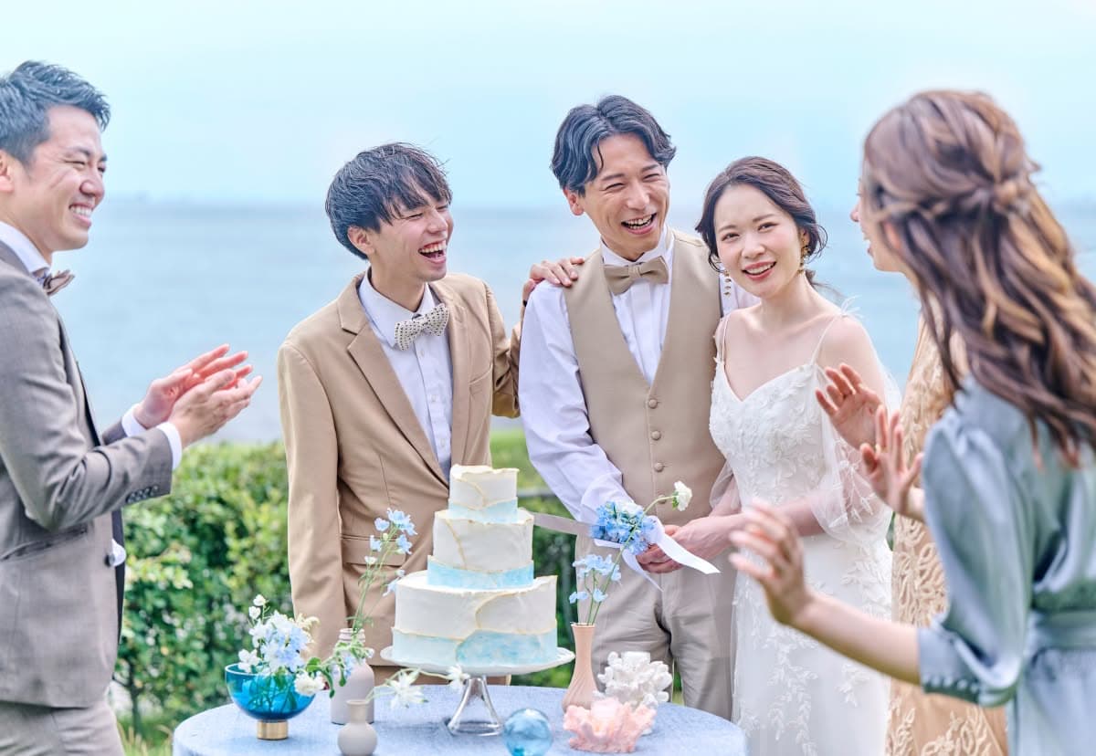 海が見える屋外会場で、新郎新婦が笑顔でケーキ入刀を行う様子。周囲ではベージュのスーツやドレスを着たゲストが拍手で祝福している。リゾートウエディングらしい、青い海と空に包まれた、和やかで幸福感あふれる披露宴のワンシーン。