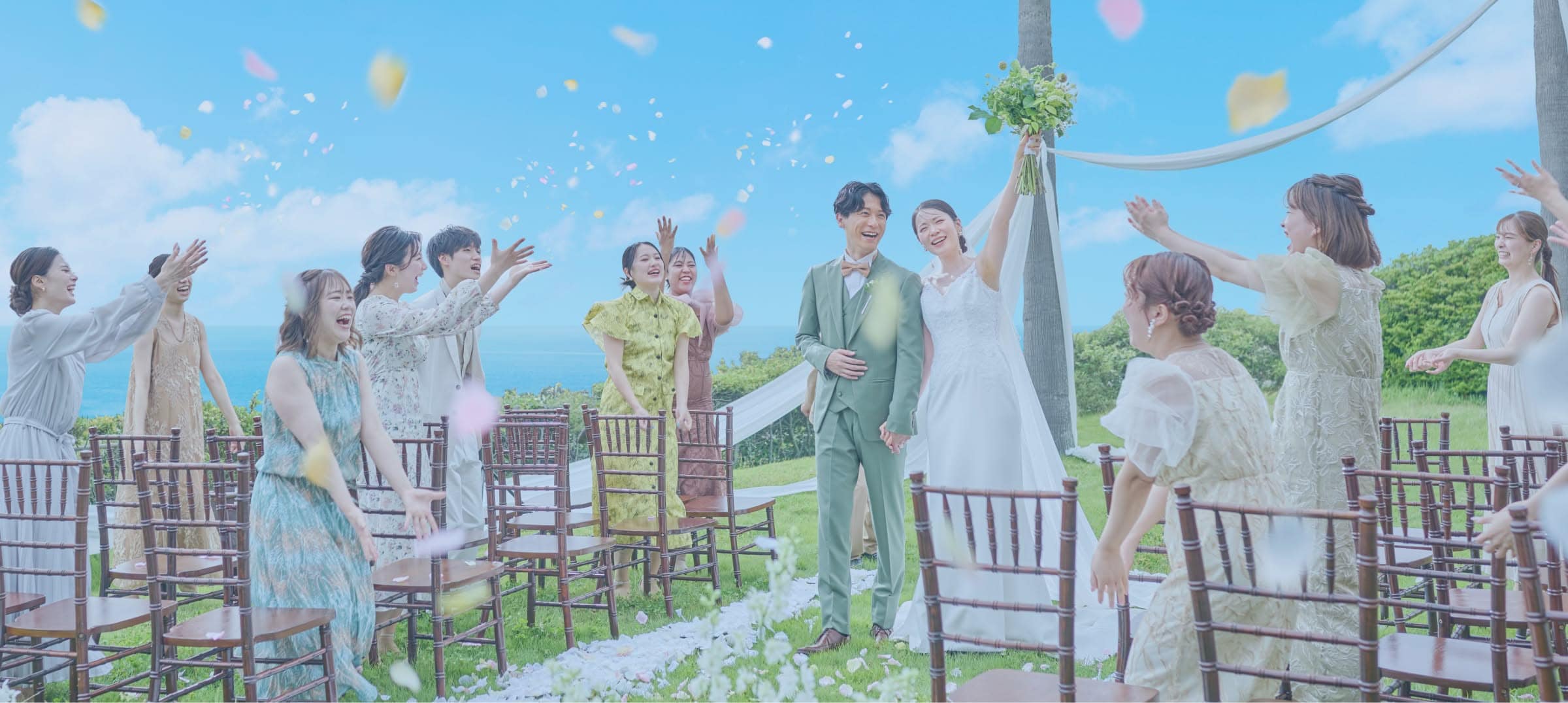 青い海を望む高台のガーデンで、ゲストからフラワーシャワーの祝福を受ける新郎新婦。新婦はブーケを高く掲げ、参列した友人たちはパステルカラーやボタニカル柄のドレスを纏い、笑顔で花びらを舞わせる多幸感あふれるワンシーン。