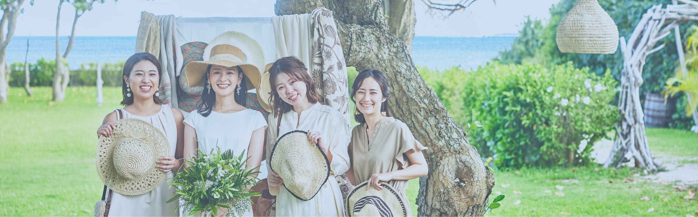 リゾートウエディングに参列する女性ゲスト4名が、屋外のガーデンで笑顔で並んでいる様子。白やベージュの軽やかなワンピースを着用し、ストローハットやブーケを手にした華やかな装い。背景には青い海と緑豊かな木々が広がる。